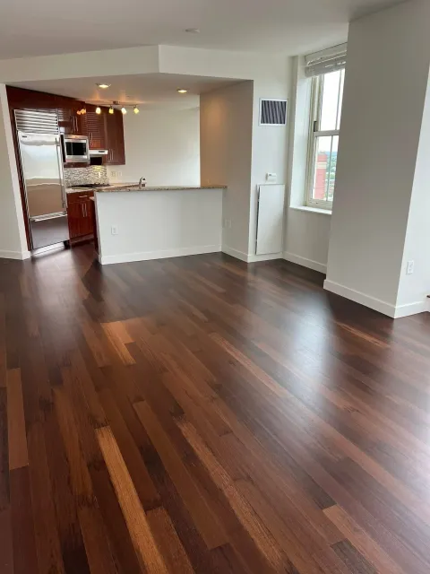 Empty living space with dark hardwood floors, white walls, and large windows letting in natural light. The partially visible kitchen has reddish-brown cabinets, a stainless steel refrigerator, and a granite countertop.