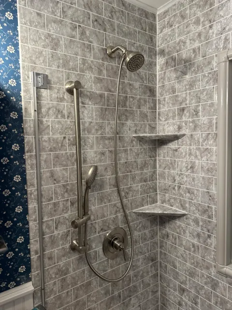 Gray tiled shower with a metal handheld showerhead, a curved metal pipe showerhead, and built-in corner shelves.