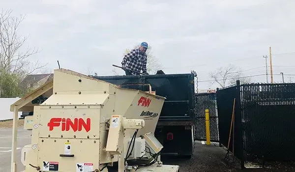 Worker atop a dumpster truck loading debris into a white FINN grinder near a fence