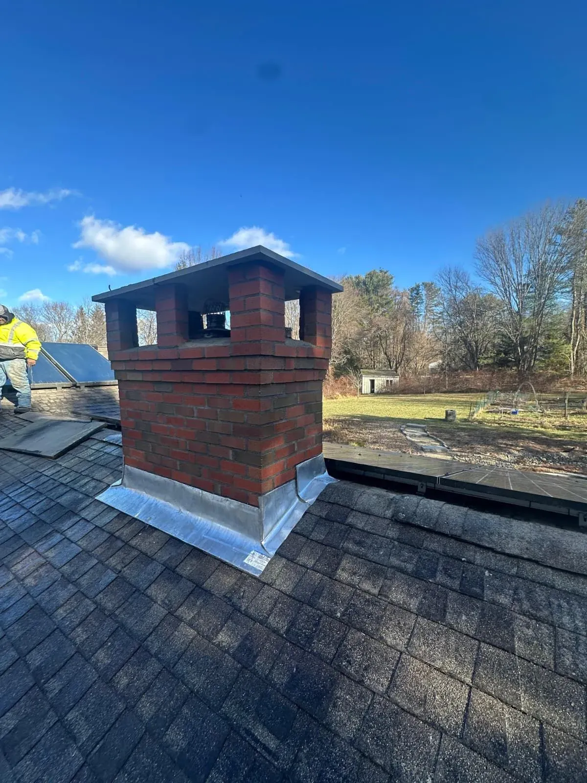 Brick chimney on a shingled roof under a blue sky, with a worker partially visible at left.