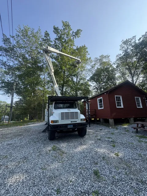 White bucket truck parked on gravel beside a red barn with the boom raised into the trees behind it