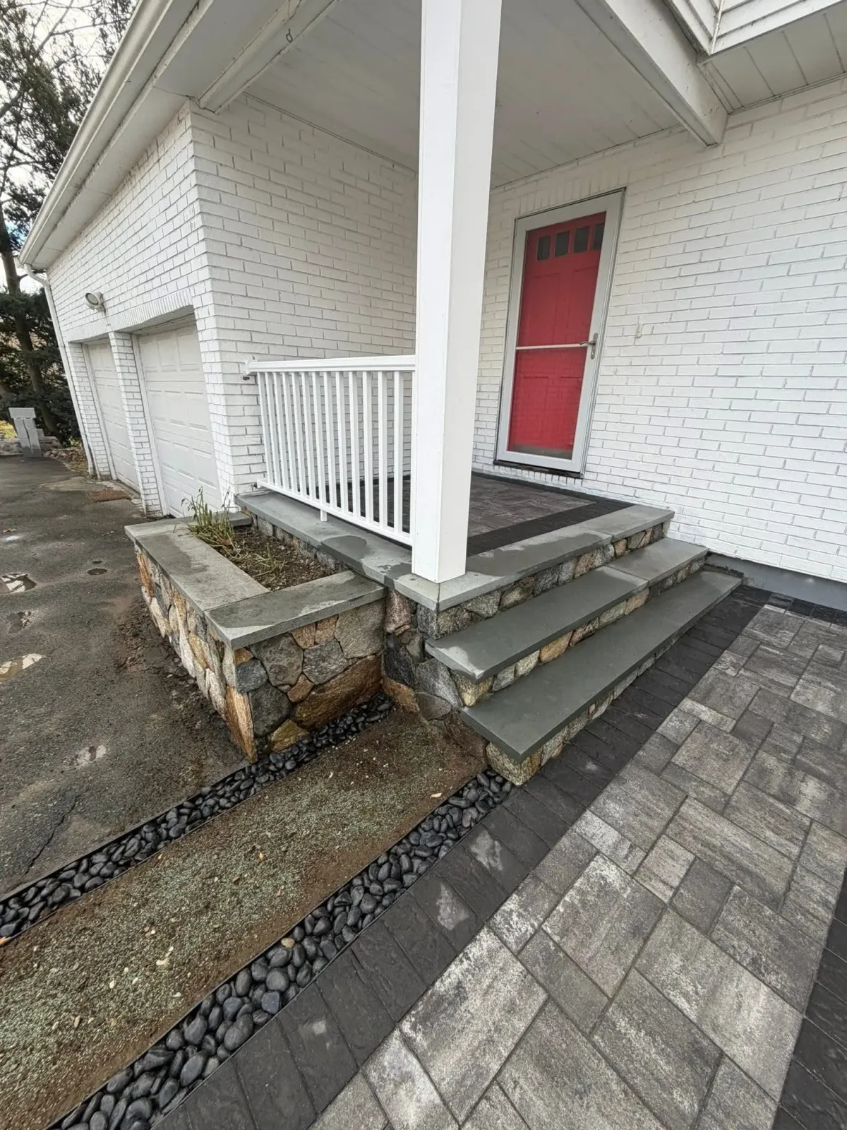 Front porch with white railing, red door, concrete steps, and paved walkway beside a white house.