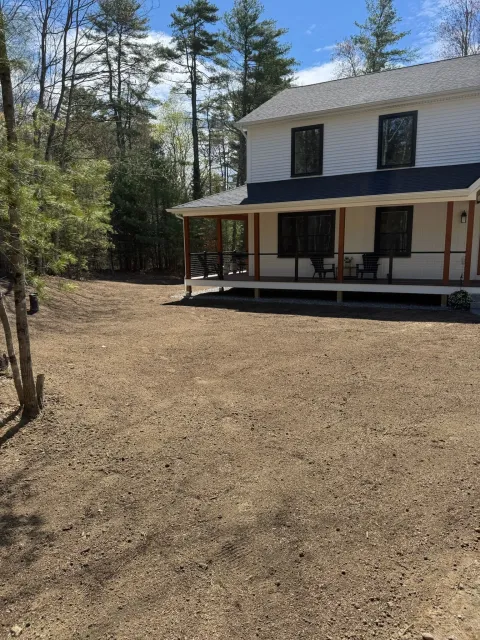 Two-story house with a covered porch beside a gravel yard and trees under a blue sky.