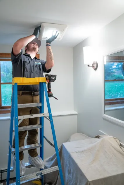 A man standing on a ladder installing or repairing a vent in the ceiling of a room with a window and a mirror on the wall.