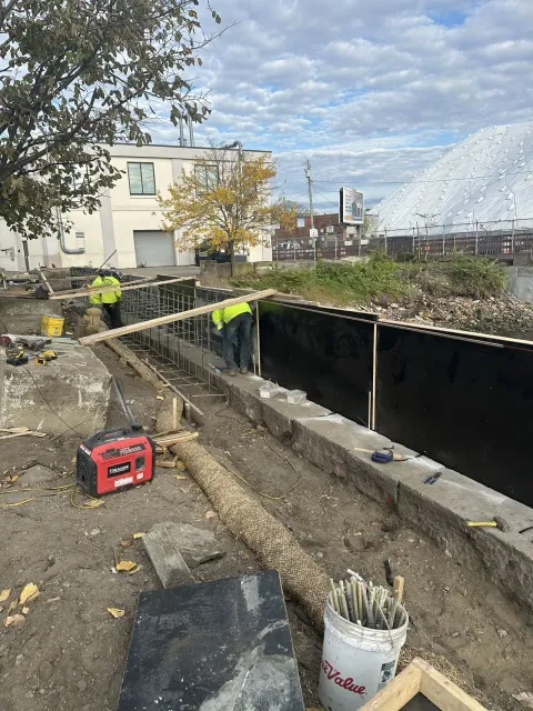 Construction workers at a site with concrete forms and black lining, working outdoors on a cloudy day.