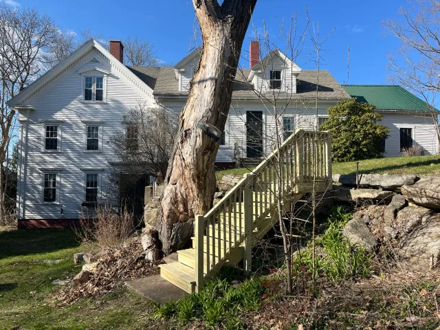 A large white house with multiple windows and a green roof, situated on a hill with rocks and trees in the yard. There is a wooden staircase leading up to the house from the yard.