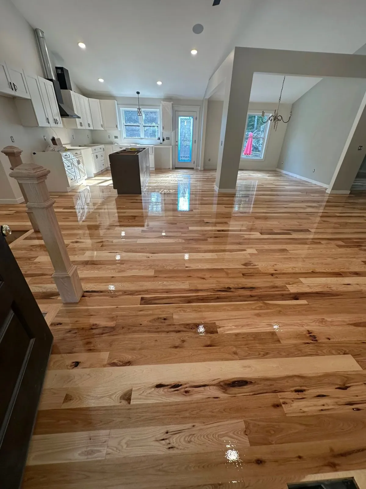 Photo of an empty kitchen with newly installed glossy wooden floors, white cabinets, and a window with sunlight shining through, including a door to the outside.