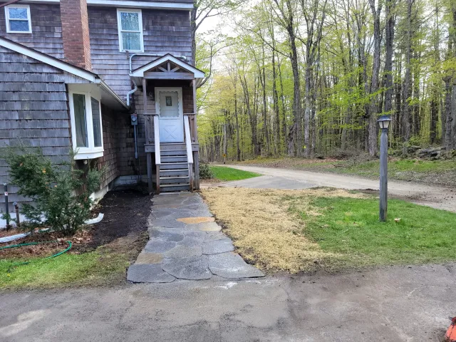 Exterior side of a house with weathered wooden siding, a small porch with stairs, and a white door. The yard has a stone pathway, a small pine tree, and a patch of dirt. There is a dirt driveway and a forest background with tall trees.