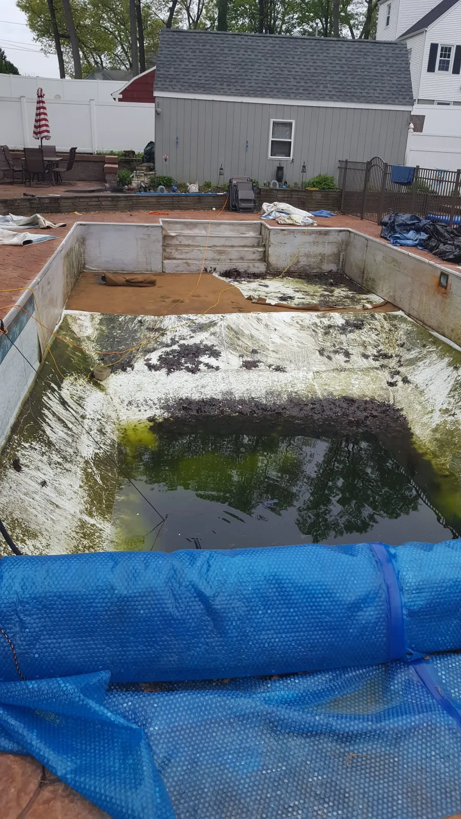 A backyard swimming pool that is empty and dirty with algae and debris, surrounded by a blue pool cover partially removed, with a fenced patio area and a small shed in the background.