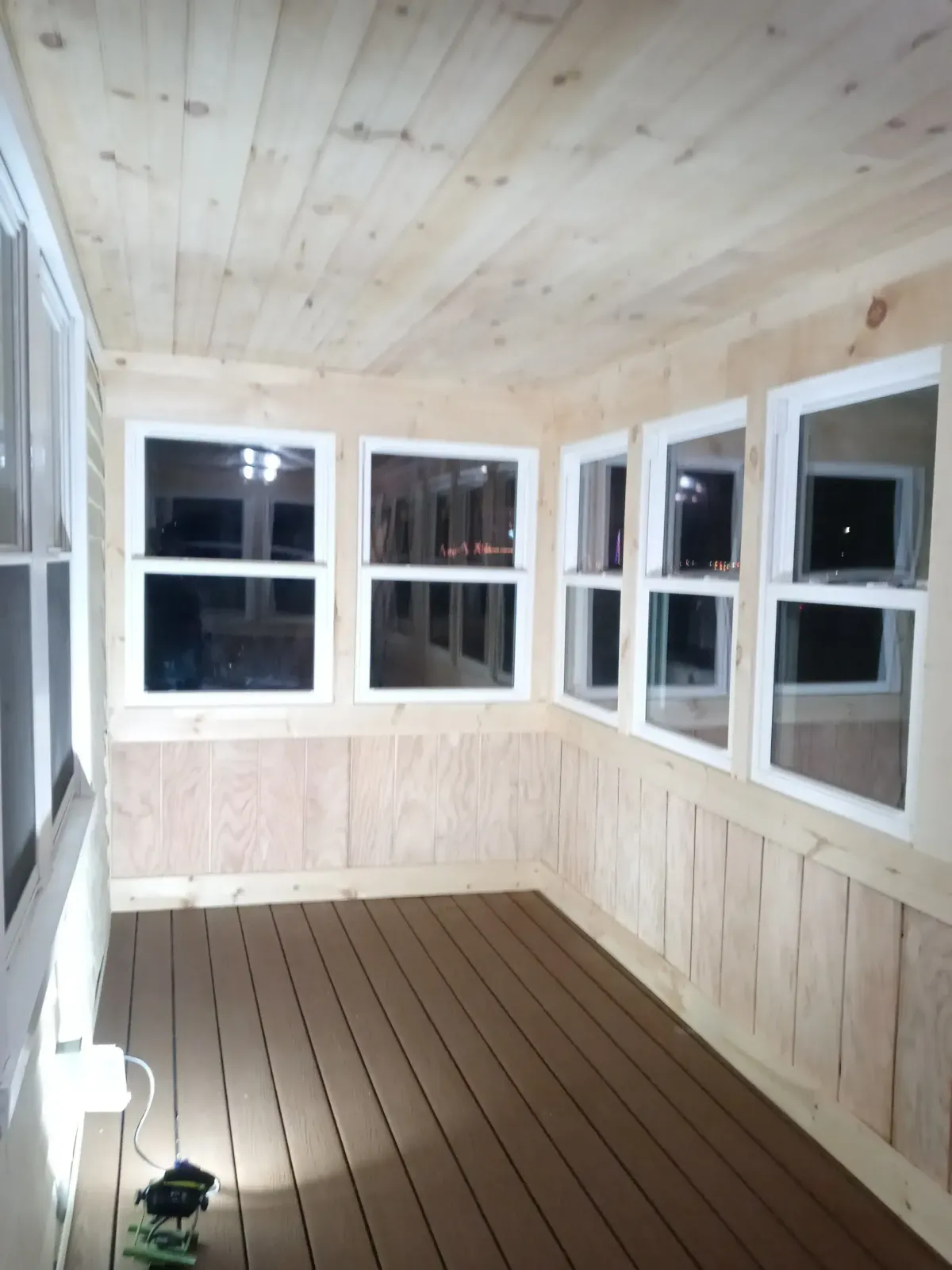 Small enclosed porch with light wood paneling, multiple windows, and a brown plank floor.