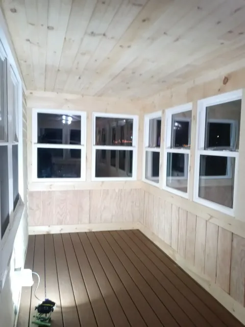 Small enclosed porch with light wood paneling, multiple windows, and a brown plank floor.