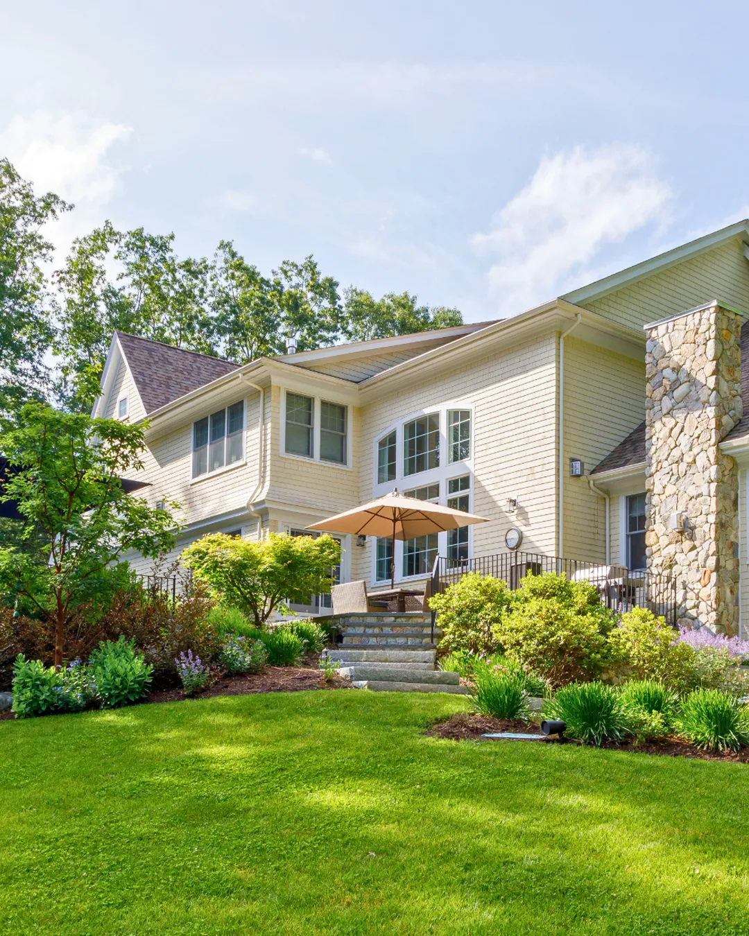A large house with light-colored siding sits amidst a lush, green lawn. The building features tall windows and a stone chimney. In front, theres a patio with an umbrella and outdoor furniture. Trees and shrubs enhance the scenic landscape.