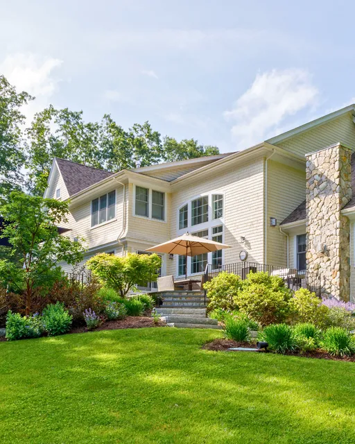 A large house with light-colored siding sits amidst a lush, green lawn. The building features tall windows and a stone chimney. In front, theres a patio with an umbrella and outdoor furniture. Trees and shrubs enhance the scenic landscape.