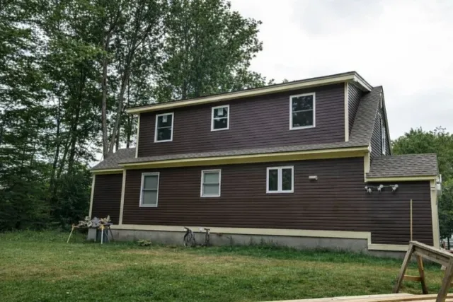 Two-story dark brown house with white-trimmed windows elevated on concrete blocks, surrounded by grass and trees
