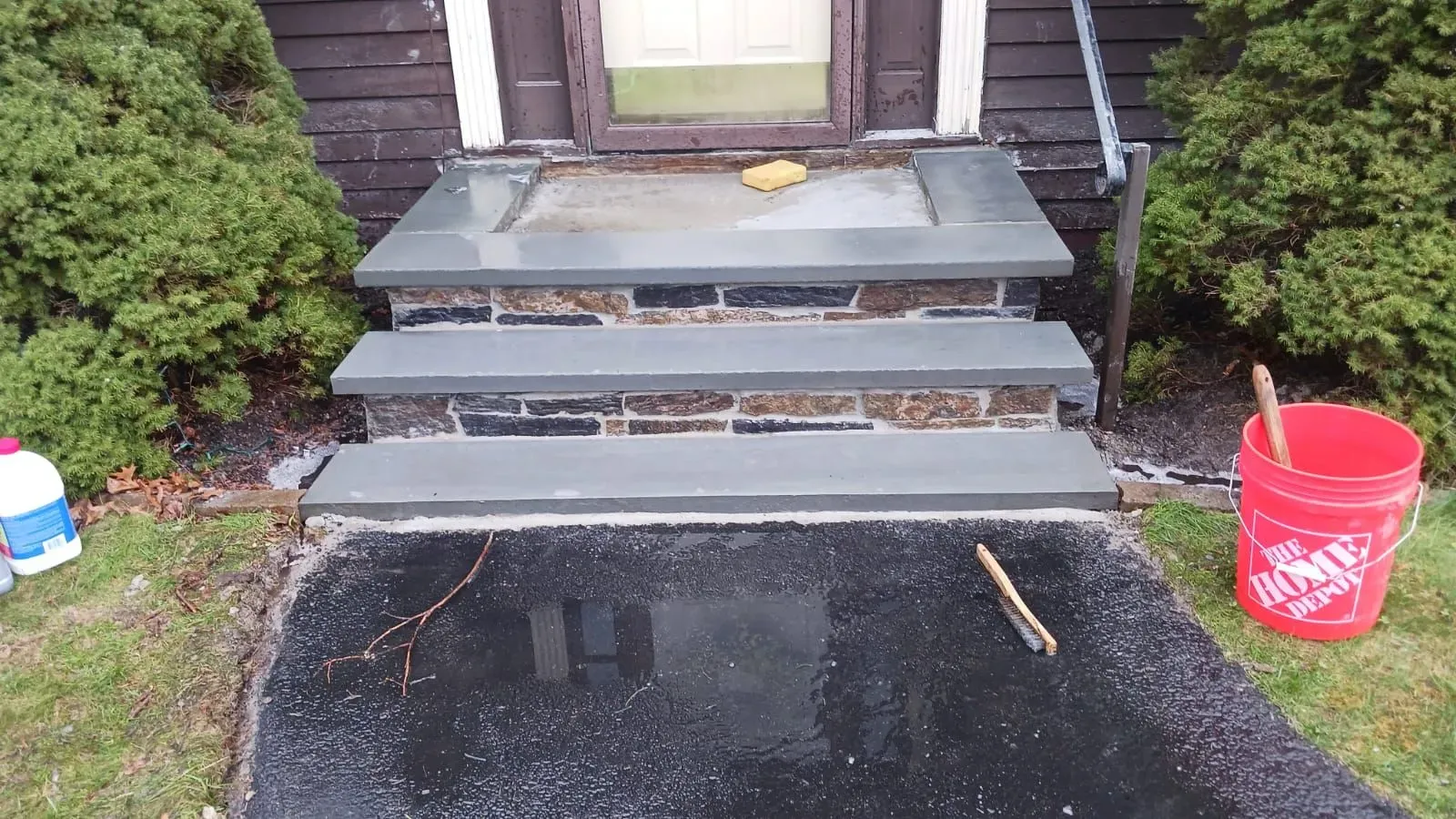 Stone steps leading to a house entrance. Wet asphalt pathway. A red bucket sits on the right.