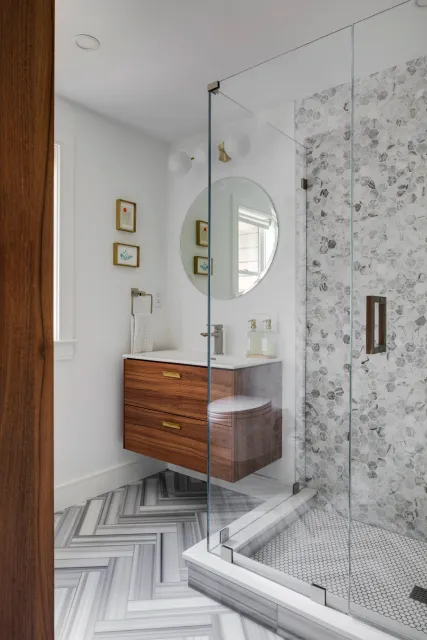 Modern bathroom with glass-enclosed shower, wooden vanity with gold handles, round mirror, small framed art, marble hex tile in shower, and chevron-patterned tile on the floor.