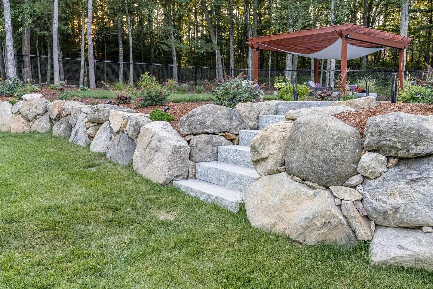 Stone retaining wall with steps leading to a wooden pergola in a backyard.