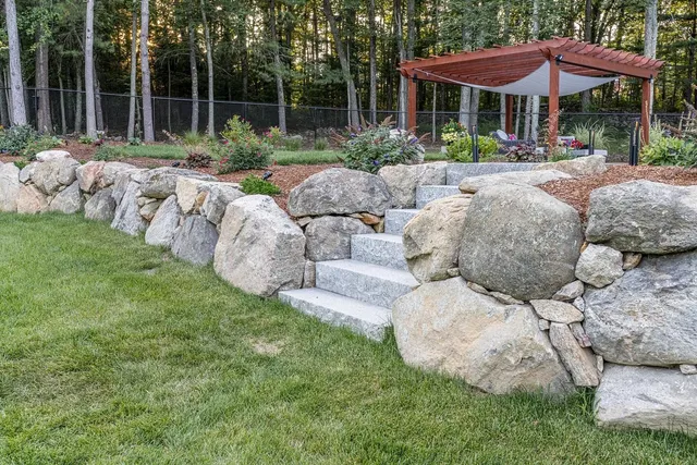 Stone retaining wall with steps leading to a wooden pergola in a backyard.