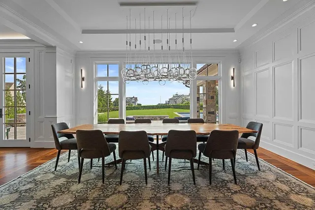 Dining room with large table, chairs, chandelier, large windows, and a view of greenery.