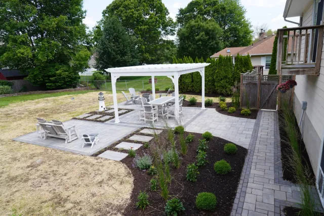 Backyard patio with white outdoor furniture, a pergola, garden beds with bushes and flowers, a lighthouse decoration, and a wooden fence with gates, surrounded by trees.