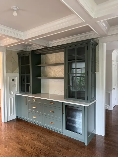 Built-in pantry cabinet in green painted millwork with glass doors, open shelving and brass hardware, set under a coffered ceiling