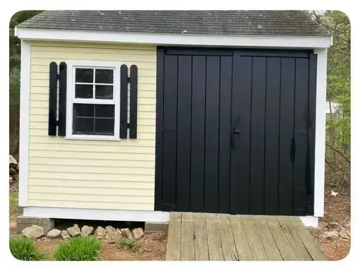 Small cream shed with black shutters and double black doors, shown with a wooden ramp.
