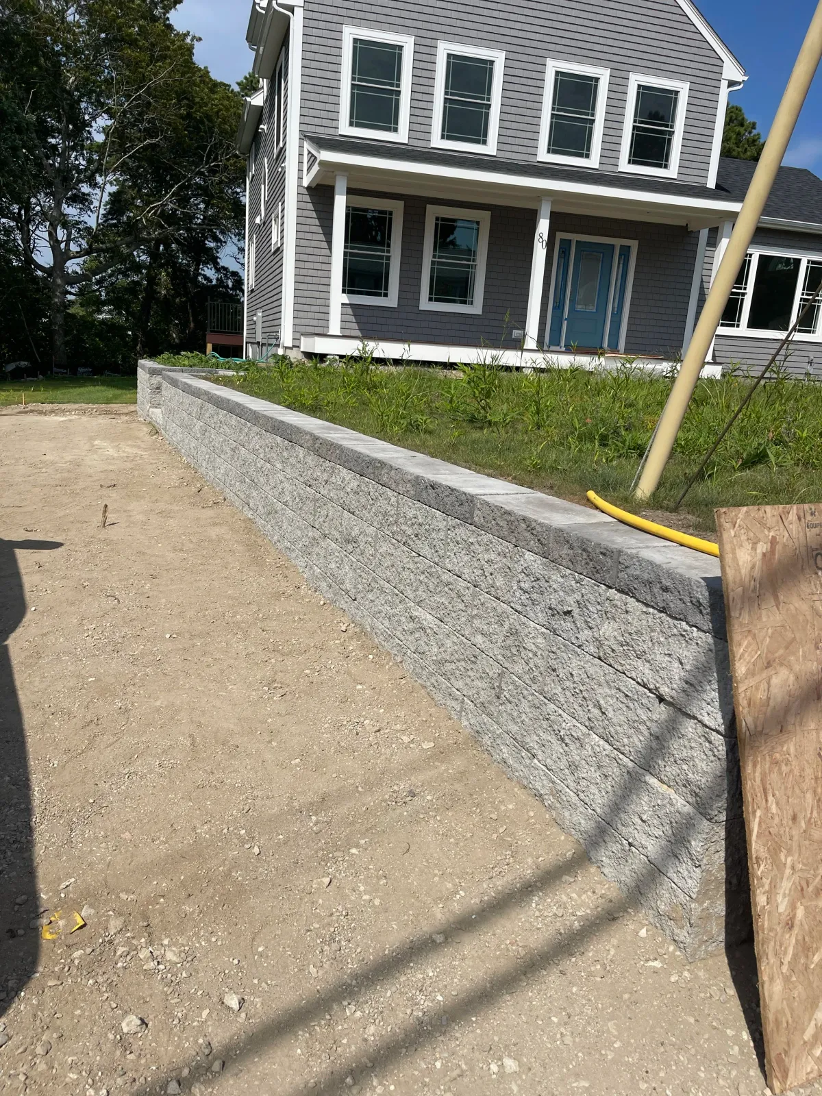 Gray house with white trim and blue door, behind a retaining wall on a dirt lot.