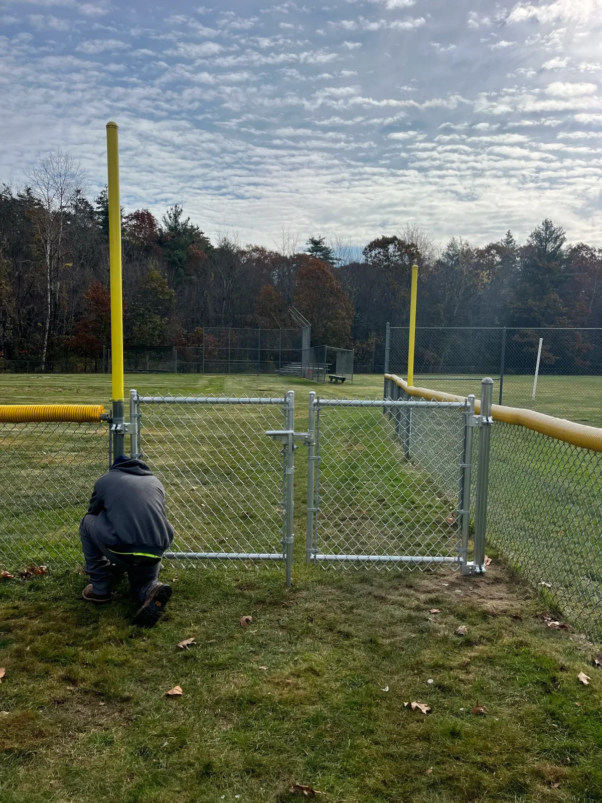Person kneeling on grass in front of a chain-link fence, approaching a yellow pole near a sports field with trees and a partly cloudy sky in the background.