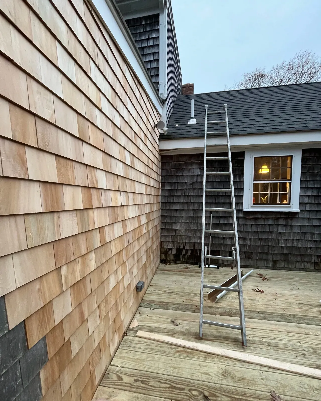 A ladder set up on a wooden deck outside a house with different siding materials, with construction tools and materials nearby.