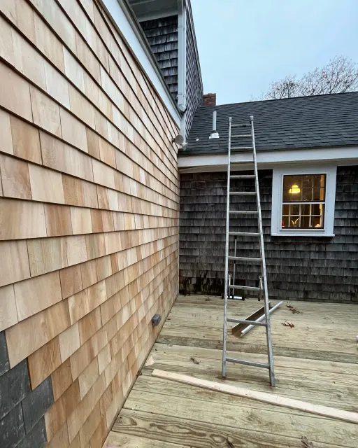 A ladder set up on a wooden deck outside a house with different siding materials, with construction tools and materials nearby.