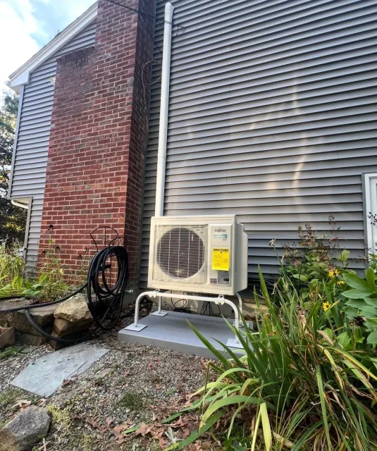 Outdoor home AC unit beside a brick wall and gray siding, with gravel and plants nearby.