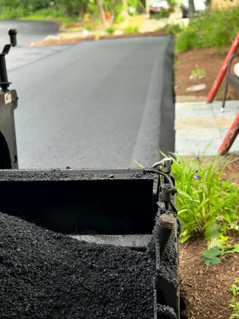 Freshly paved asphalt road alongside a garden bed; a truck bed filled with asphalt in foreground.