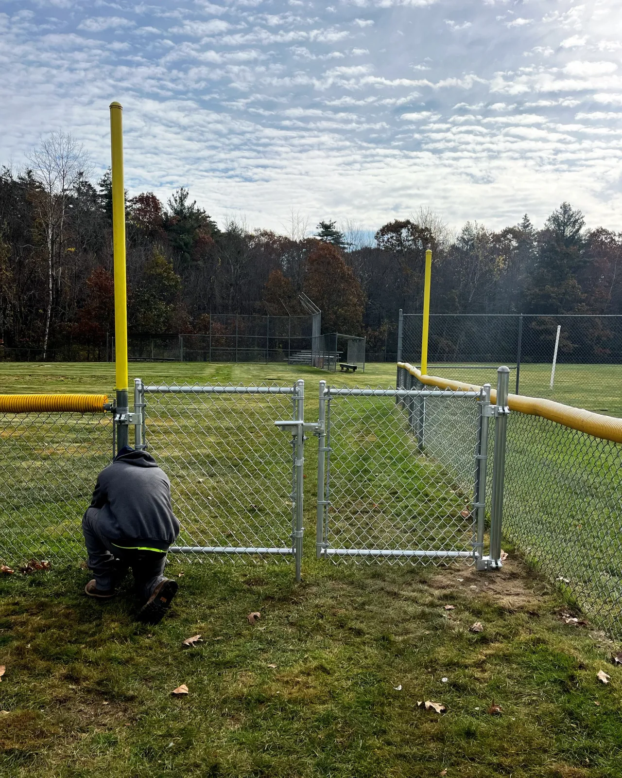 A person kneeling on the grass next to a chain-link fence at a sports field, with yellow goalposts and trees in the background under a partly cloudy sky.