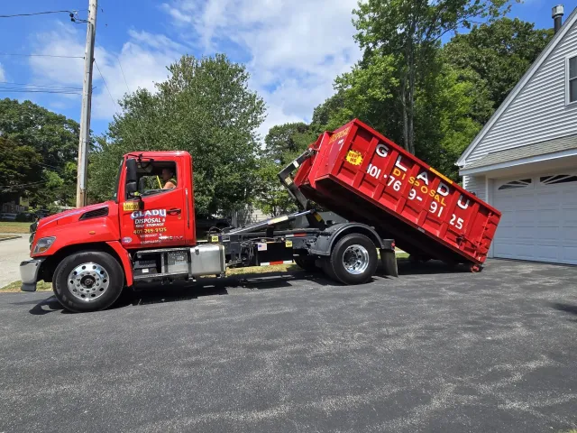 A red dumpster truck parked on a driveway with the dumpster slightly elevated at an angle. The truck has company branding, including a phone number, and is set in a residential neighborhood with trees, houses, and a partly cloudy sky in the background.