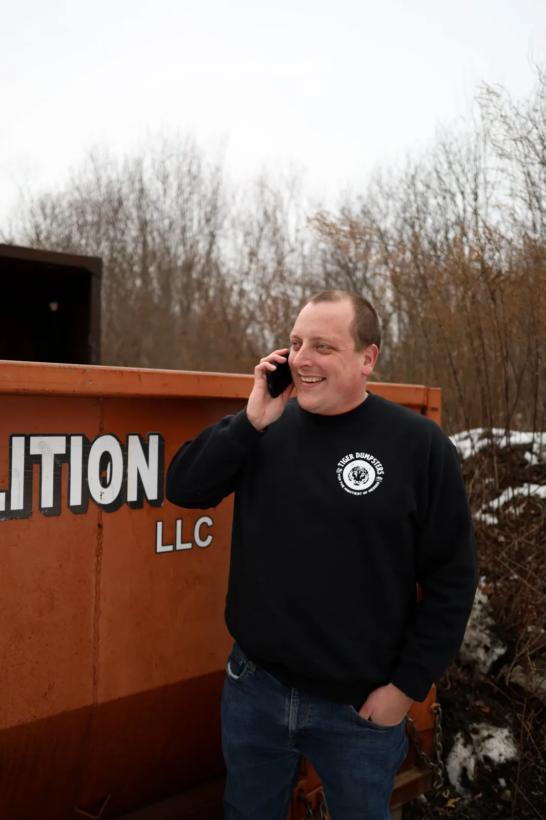 A man smiling and talking on a cell phone outdoors next to an orange dumpster.