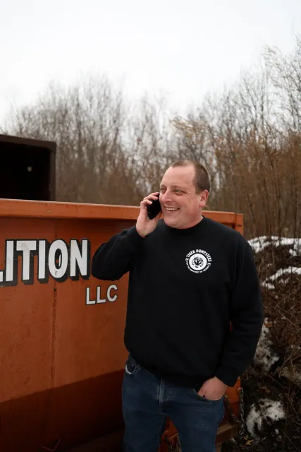 A man smiling and talking on a cell phone outdoors next to an orange dumpster.
