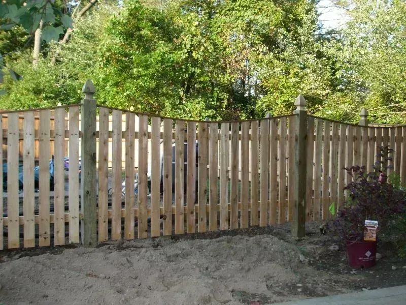 A newly installed wooden picket fence with a scalloped top edge, supported by sturdy posts, standing before green trees.