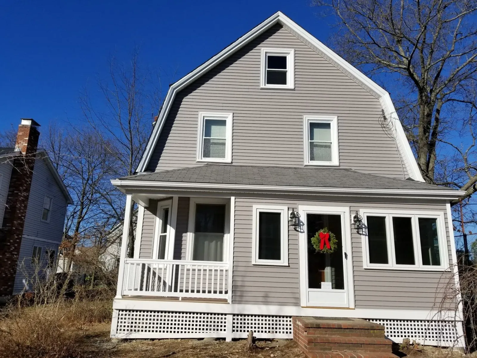 Gray house with a porch and a red wreath on the door; blue sky in the background.