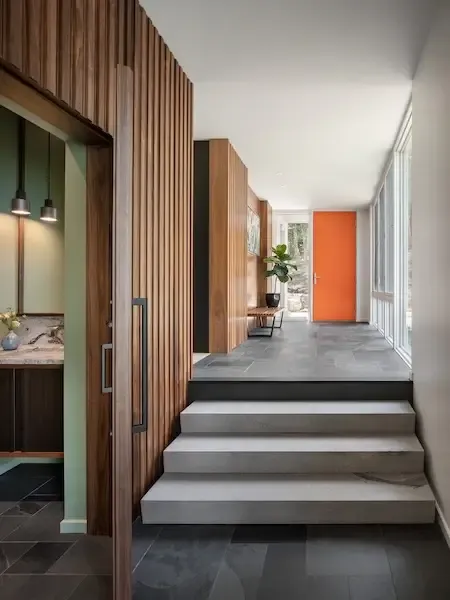 A modern hallway by a Maine architect features wood paneling, gray tile floors, white steps, and a bold orange door. Large windows on the right flood the space with natural light.