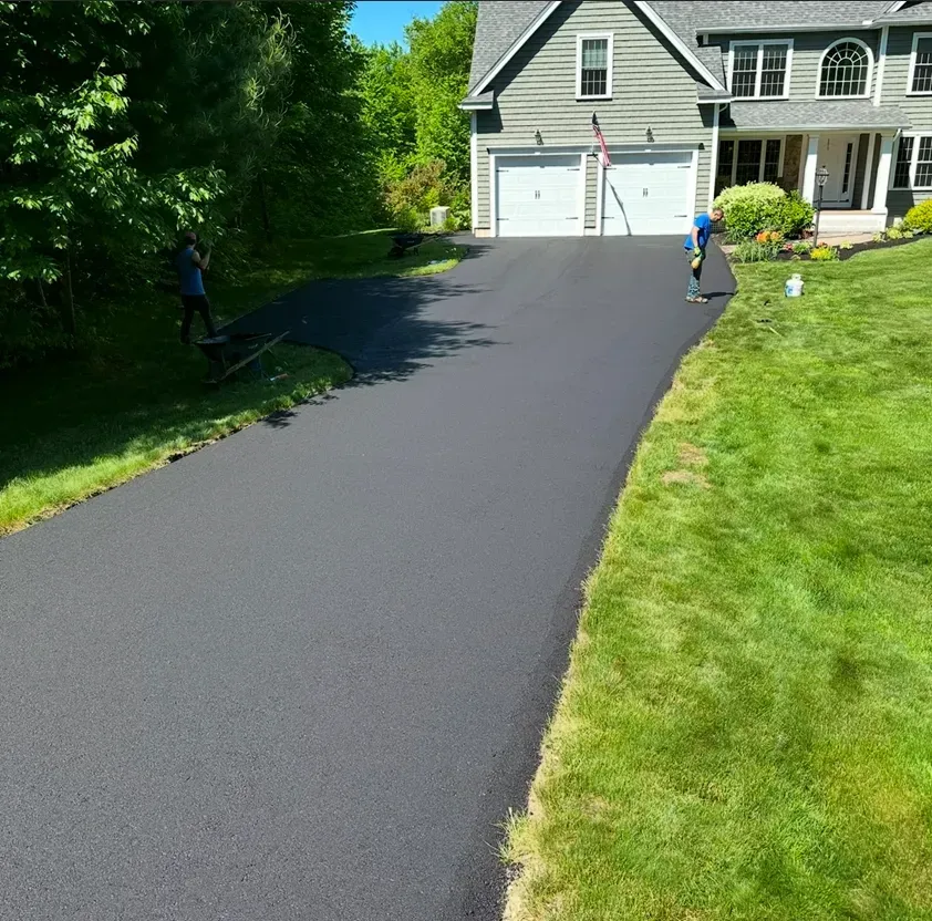 A freshly paved black asphalt driveway leads to a gray suburban home with two garage doors on a sunny day.