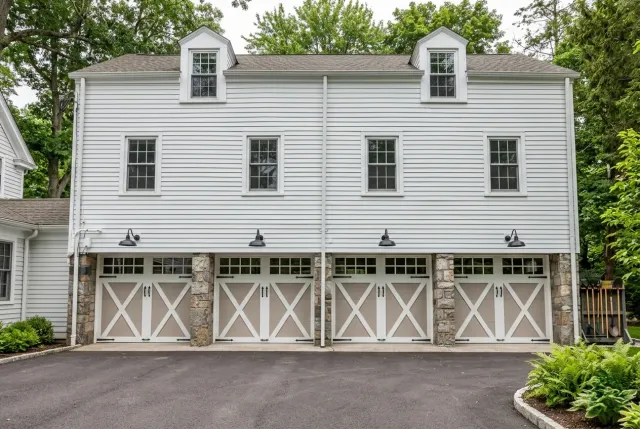 White two-story carriage house with four garage doors, dormer windows, and a paved driveway