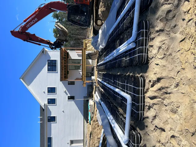 Hitachi excavator grading dirt site next to new metal-sided commercial building with blue drainage pipes