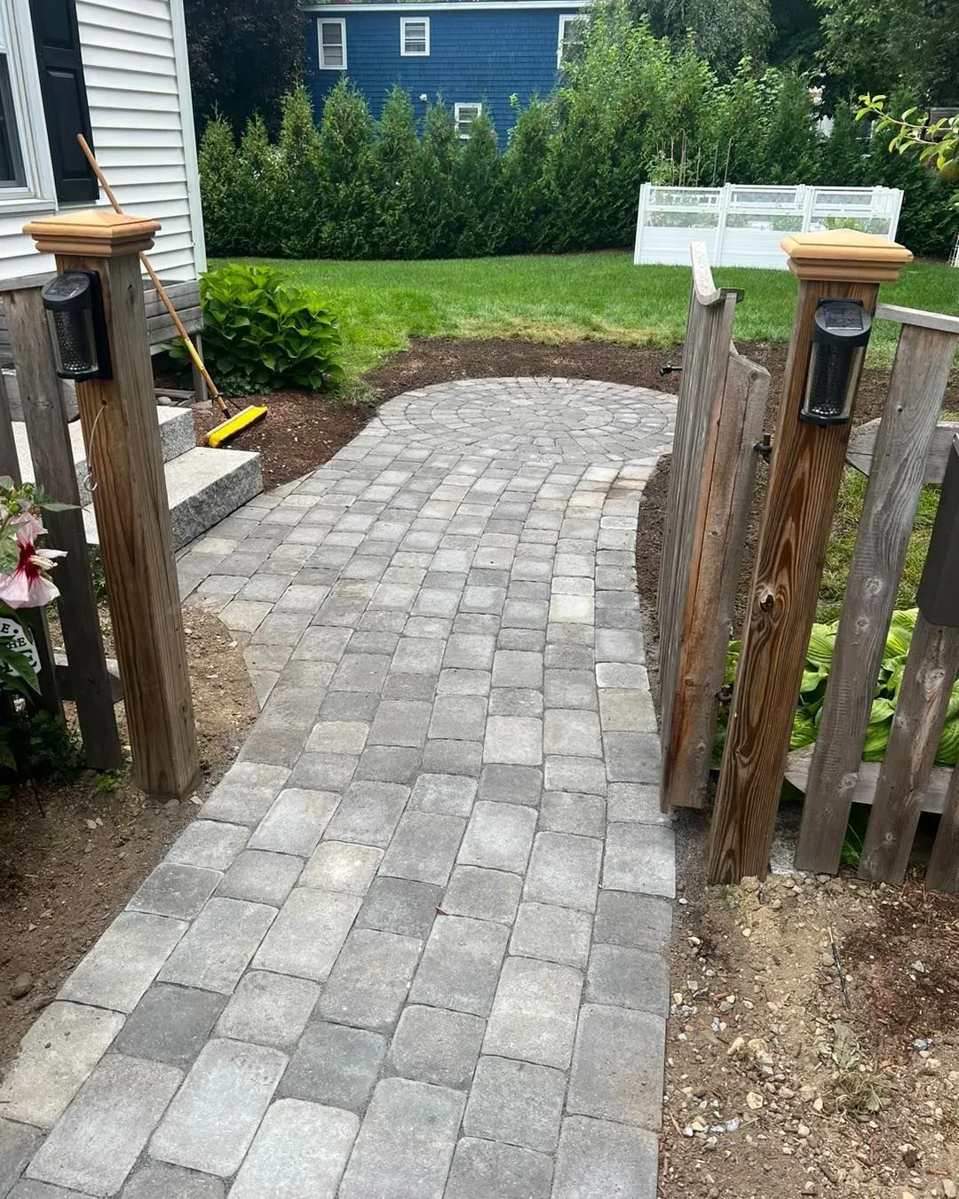 A gray paver walkway leads through a wooden fence opening towards a green lawn and a blue house.