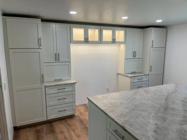 A kitchen with white cabinets, some with frosted glass doors, and marble countertops. Recessed ceiling lights illuminate the space, and there is wooden flooring.