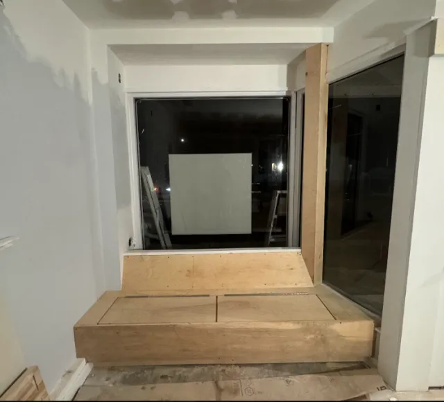Interior of a room under construction, with a window and a sliding glass door, and unfinished wood platform and framing.
