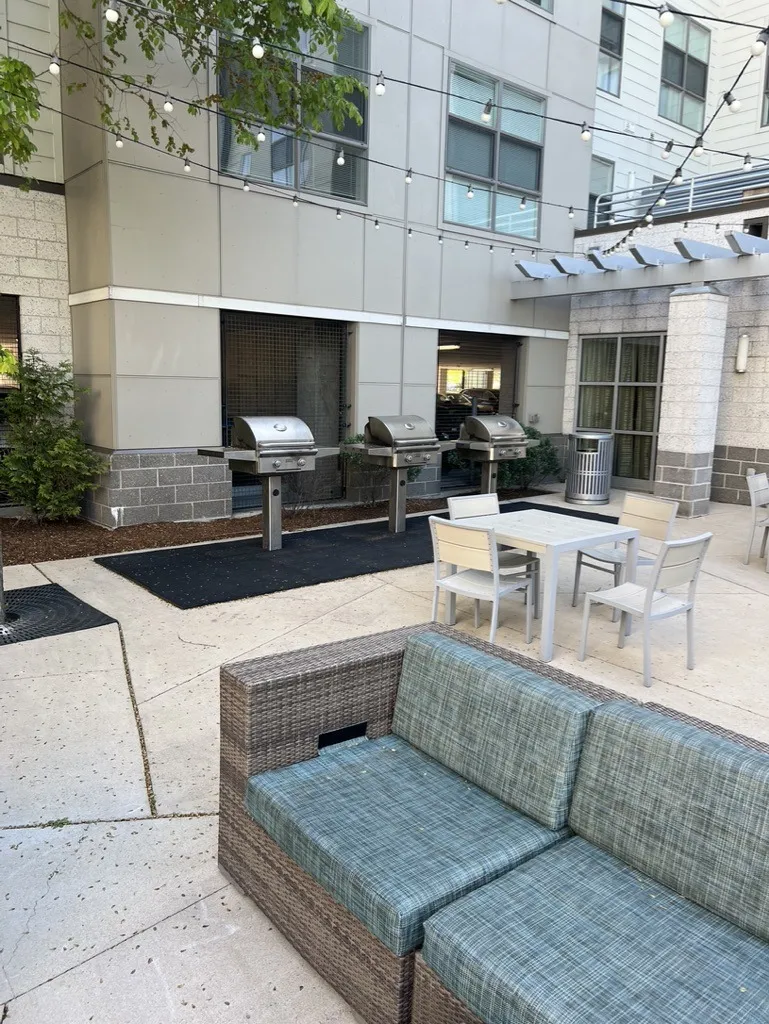Outdoor patio with three grills, seating area, and string lights. Modern building facade in background, surrounded by greenery and potted plants.