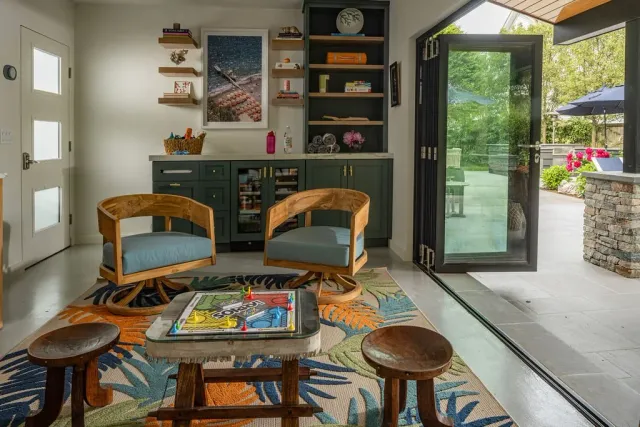 Cozy indoor space with two wooden chairs with light blue cushions, a colorful leaf-patterned rug, and an open glass door leading to a patio with blooming pink flowers and greenery.