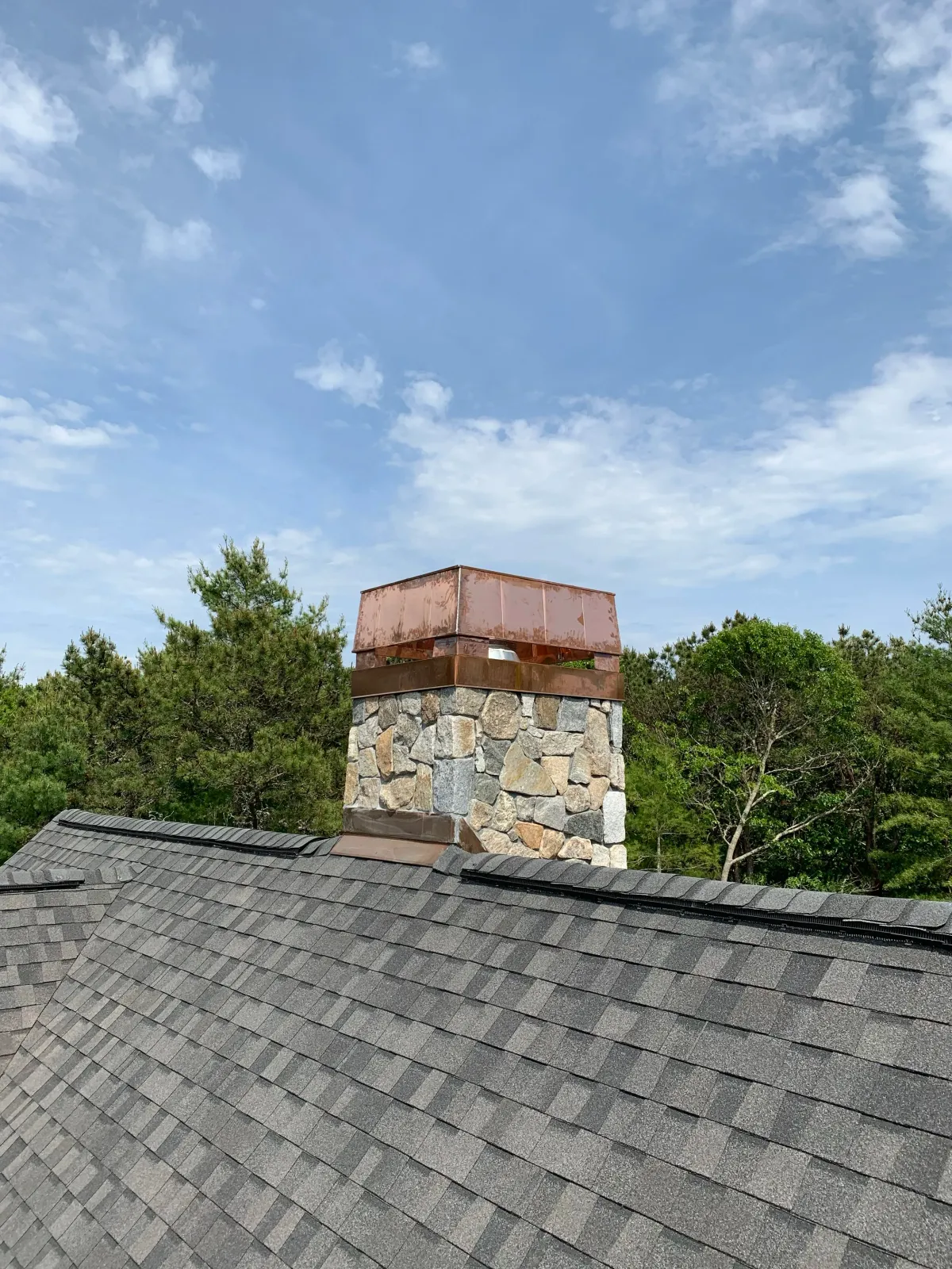 A rooftop with gray shingles and a stone chimney topped with a rusted metal cap, against a blue sky with scattered clouds and green trees in the background.