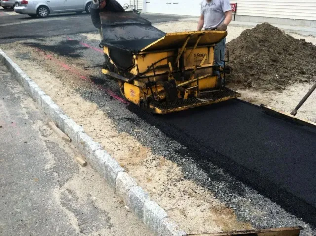 Asphalt paving machine laying blacktop on a prepared gravel bed, curb on the left, worker guiding the machine.