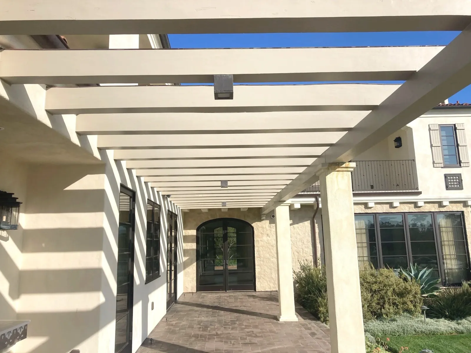 View of a covered patio with white pergola casting striped shadows, black-framed glass doors and windows, stone columns, and landscaped bushes and plants.